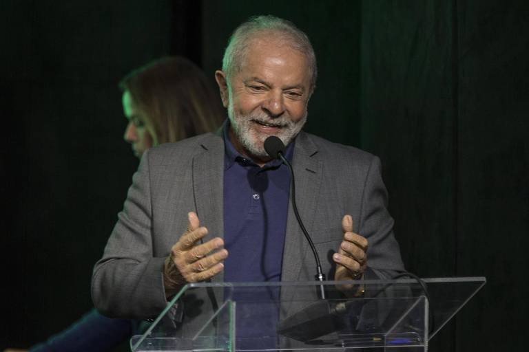 Lula está atrás de microfone e sorri. Ele veste um blazer cinza e gesticula com as duas mãos