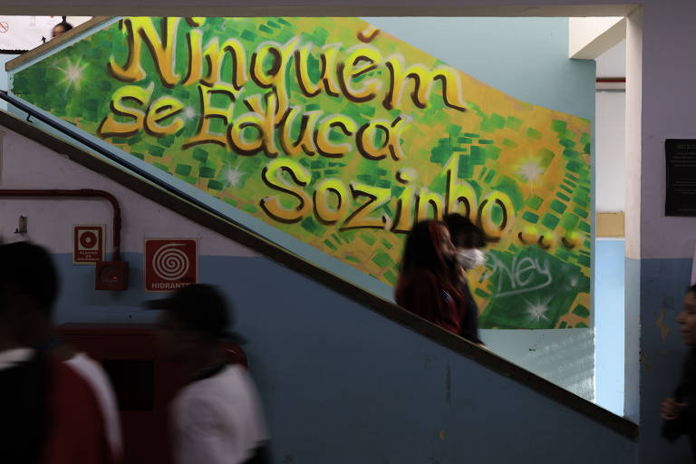 Imagem mostra um corredor de uma escola da capital paulista. Alguns estudantes passam por um mural onde se lê: "ninguém se educa sozinho". O mural mescla as cores verde e amarelo.