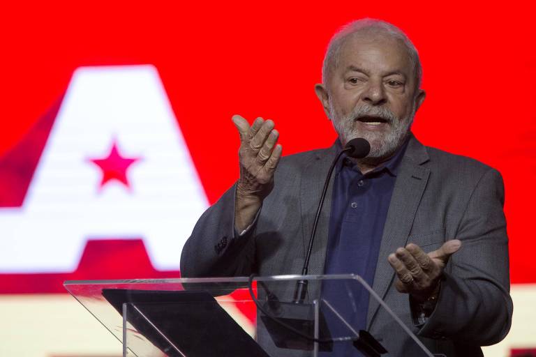 Lula veste blazer cinza e gesticula com as duas mãos. Ele fala atrás de microfone. Atrás, há um painel com as cores vermelha e branca