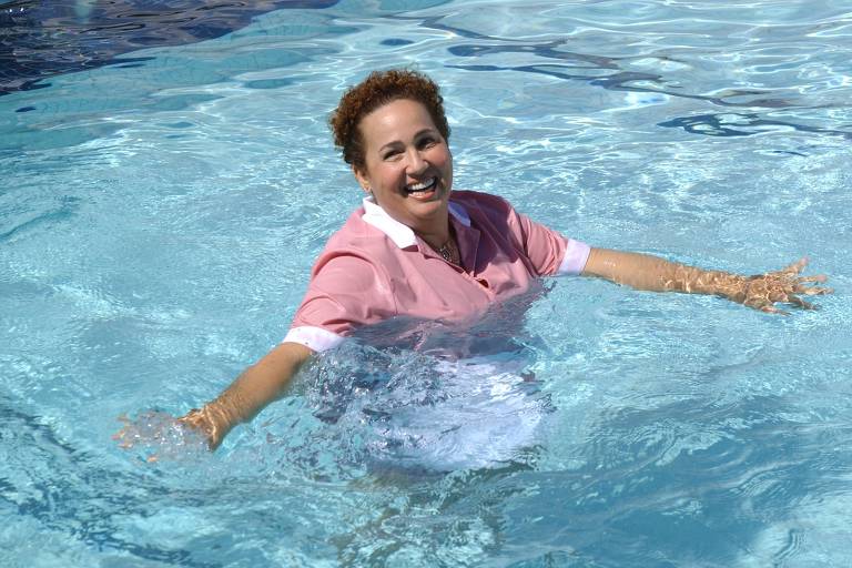 Uma mulher com uniforme de empregada doméstica está na piscina, com metade do corpo submersa, ela ri para a câmera