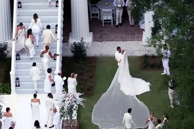 Em foto colorida, noivos se beijam diante dos convidados em uma cerimônia no jardim de uma casa
