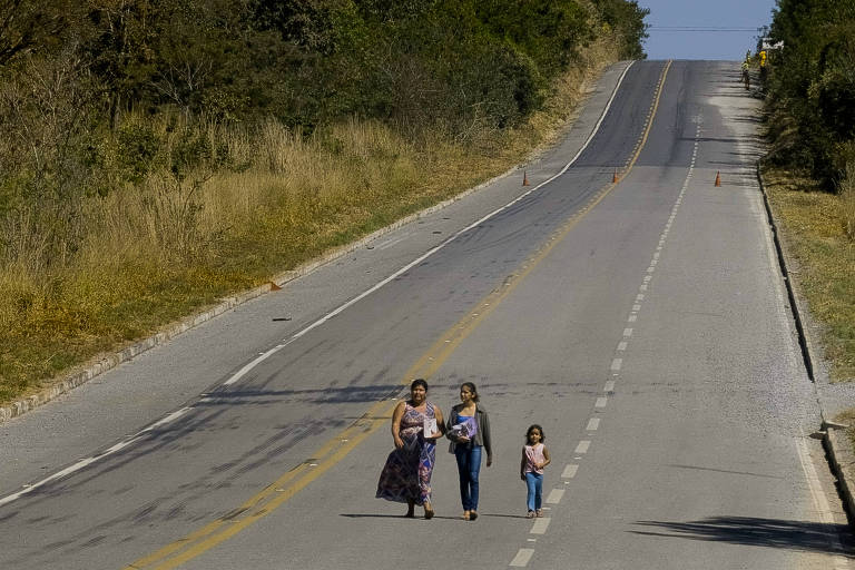 Imagem geral mostra extensão de uma rodovia, sem movimento de veículos, com 3 pessoas caminhando no meio da pista: 1 mulher adulta, acompanhada de uma adolescente e uma menina. Elas caminham na longa estrada esvaziada, com vegetações nas laterais. 