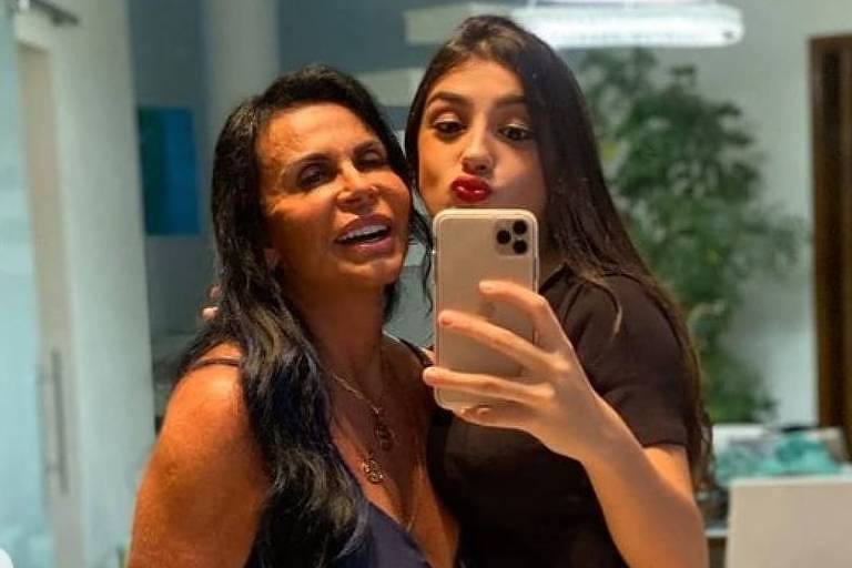Em foto colorida, duas mulheres com vestidos pretos jogam beijos para a câmera enquanto fazem selfie