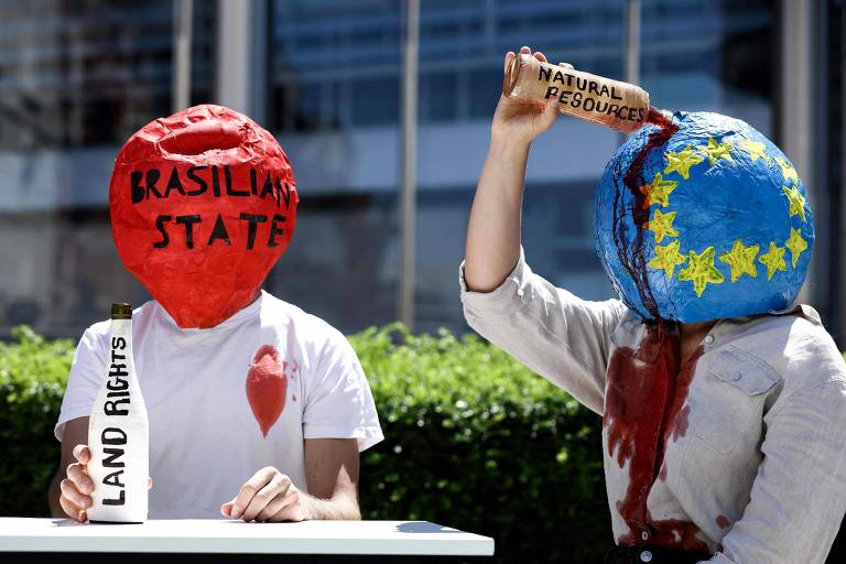 Pessoas com globos na cabeça, um representando o Estado brasileiro e outro a União Europeia, cravam garrafas nessas estruturas e um líquido vermelho, como se fosse sangue, escorre por suas roupas
