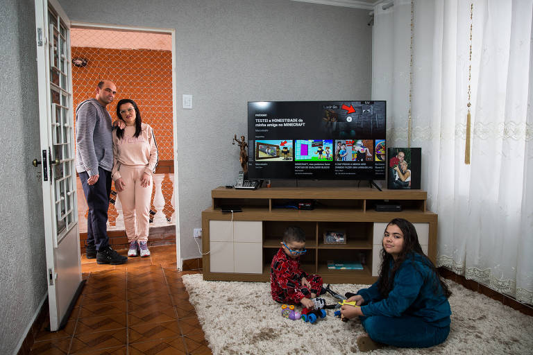 Casal branco, de pé, próximo a duas crianças sentadas no chão sobre um tapete felpudo, diante de uma TV.