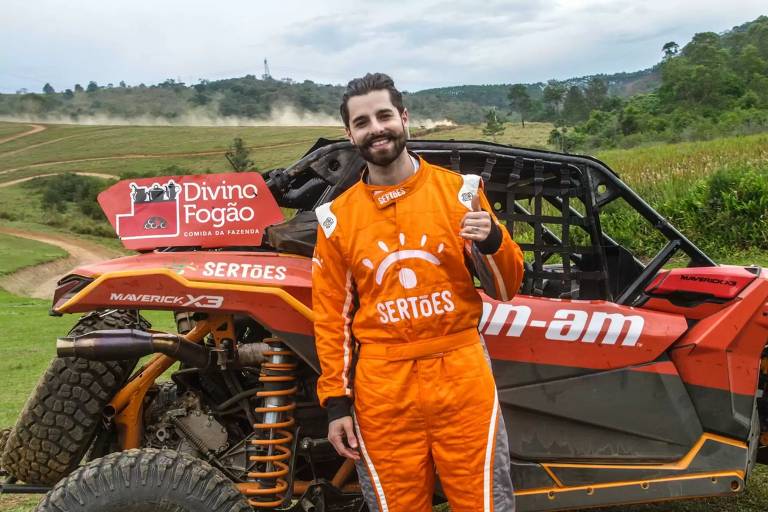 O DJ Alok, branco de barba e vestindo um macacão de corrida laranja posa em frente a um carro do Rally dos Sertões