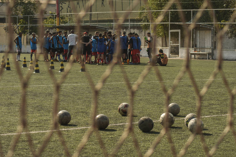 Imagem mostra um campo de futebol através da rede de um gol. Há várias bolas no chão e, no fundo, um grupo de meninos reunidos em uma roda.