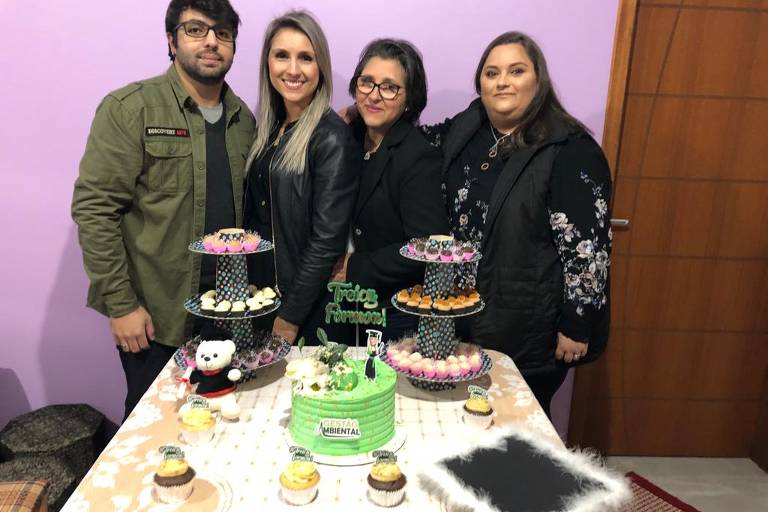 Mãe e três filhos, sendo um homem e duas mulheres, estão reunidos em volta de uma mesa com bolo e enfeites de aniversário