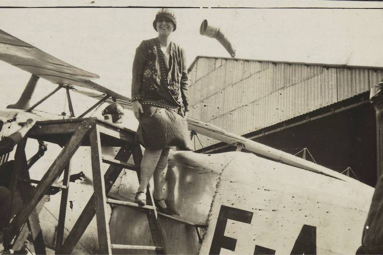 Uma mulher de vestido na altura do joelho, salto e chapéu, com cabelos curtos, se equilibra em uma escada, prestes a subir em um avião monomotor antigo