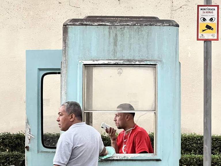 A imagem mostra uma cabine azul em um ambiente urbano. Dentro da cabine, há um homem vestido com uma camiseta vermelha e um celular na mão próximo à boca. Do lado de fora, um homem de camisa cinza está apoiado na janela da cabine. Ao fundo, há uma parede clara e uma cerca viva com plantas. Há uma placa com desenhos de olhos, de uma câmera de segurança e o aviso "monitorada 24 horas".
