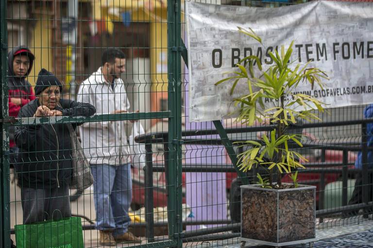 Idosa é vista atrás das grades de um portão, junto a outras pessoas, ao lado de uma faixa escrita 'O Brasil tem fome' 