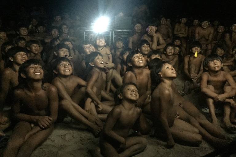 Crianças indígenas sentadas no chão observam uma projeção; ao fundo, é possível ver a luz do projetor