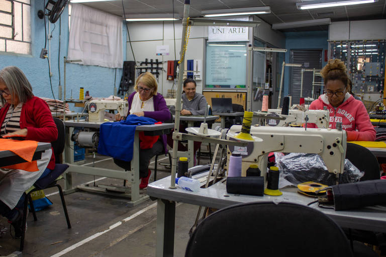 Imagem colorida mostra três mulheres à esquerda e uma à direita; elas estão sentadas, trabalhando em máquinas de costura, dentro de um galpão