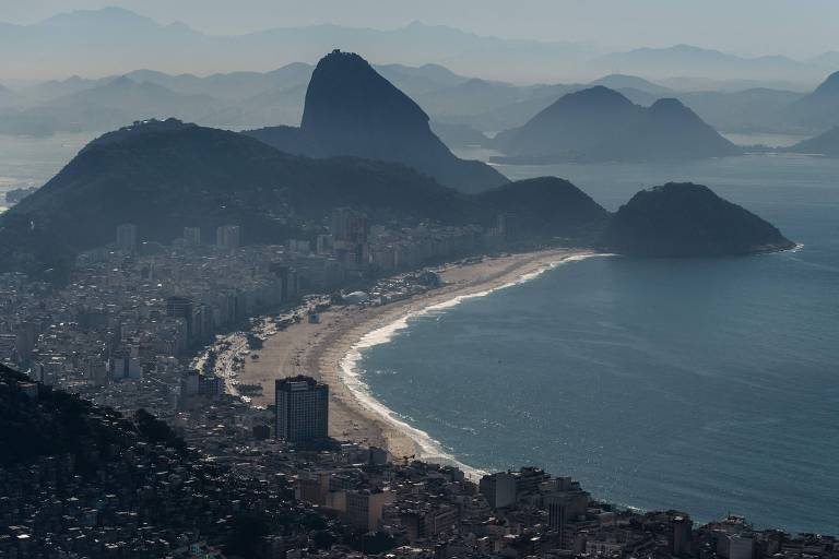 Visão aérea da praia de Copacabana. Em terra, há vários prédios. No fundo da imagem, é possível ver vários morros, entre eles o Pão de Açúcar.