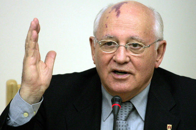 Fotografia colorida do ex-líder soviético Mikhail Gorbatchov em plano médio. Ele é um homem branco, calvo, com cabelos brancos na lateral da cabeça; tem olhos castanhos e usa óculos ovais de aros metálicos, paletó preto e camisa e gravata cinzas, enquanto fala em um microfone com a mão esquerda levantada