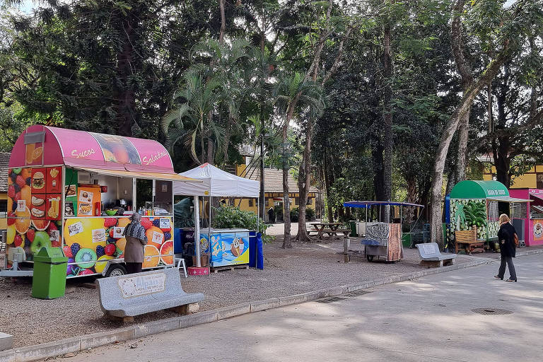 Imagem de três trailers de lanches, bebidas, sorvete e água de coco, e um carrinho de pipoca no parque da Água Branca. Na frente deles há alguns bancos vazio e, ao fundo, as árvores do parque.