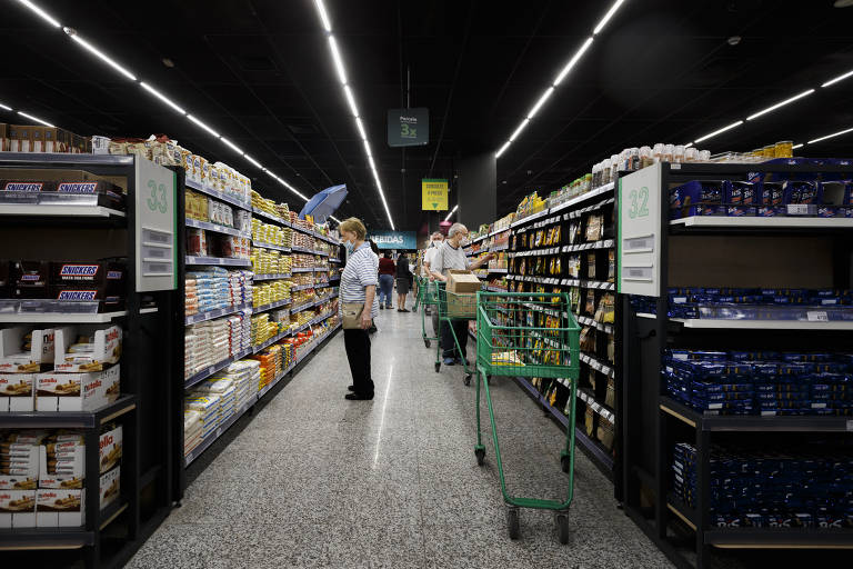 Mulher branca e aparentemente idosa está parada em pé olhando prateleiras de um supermercado. Ela está ao fundo da imagem e o há duas pessoas próximas a ela. Mais a frente, há um carrinho de supermercado verde