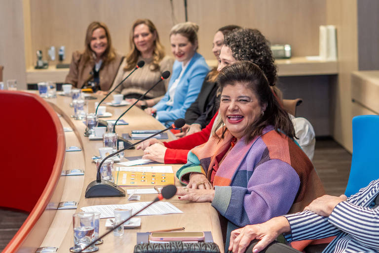 Seis mulheres sentadas em uma mesa longa, com microfones à frente