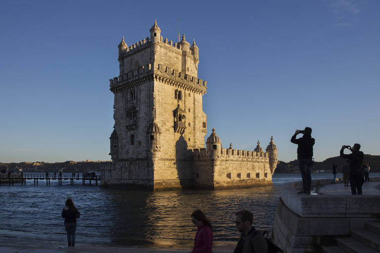 Imagem da Torre de Belém, uma construção manuelina em cores claras, em meio às águas do rio Tejo. A foto tem ainda alguns turistas em torno do ponto turístico