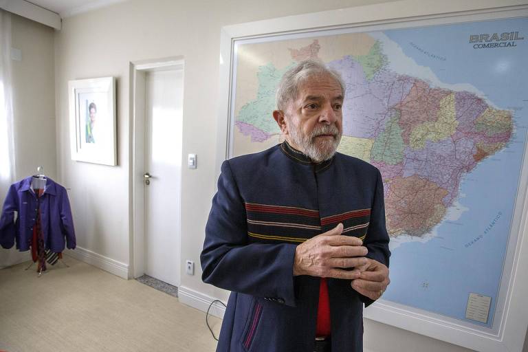 Lula, homem branco, veste jaqueta escura com listras; ao fundo, quadro grande com mapa do Brasil e porta