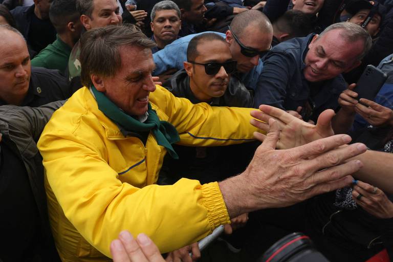 Jair Bolsonaro é um homem branco, idoso, cabelo preto com mechas grisalhas, sem barba, nariz pontudo. Está vestido com uma blusa verde e amarela, nas cores da bandeira nacional. Na foto, está no centro, à esquerda. Com a mão direita levantada, cumprimenta os apoiadores, que estão ao redor em toda a imagem