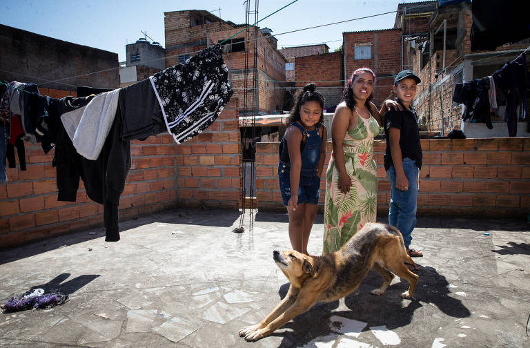 A imagem mostra uma mulher e duas crianças com o cachorro vira-latas de estimação; eles estão num quintal ensolarado na comunidade onde moram