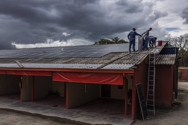 Imagem mostra painéis solares em um telhado. Dois operários estão próximos aos painéis.