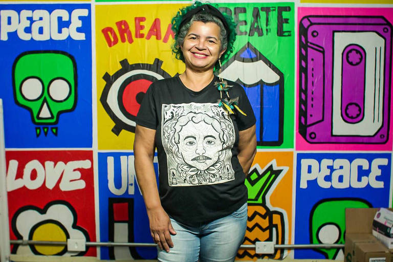 Retrato de mulher com camiseta com ilustração do rosto de Chico Mendes; ela sorri; ao fundo há cartazes de cores vivas com palavras como "peace" (paz)