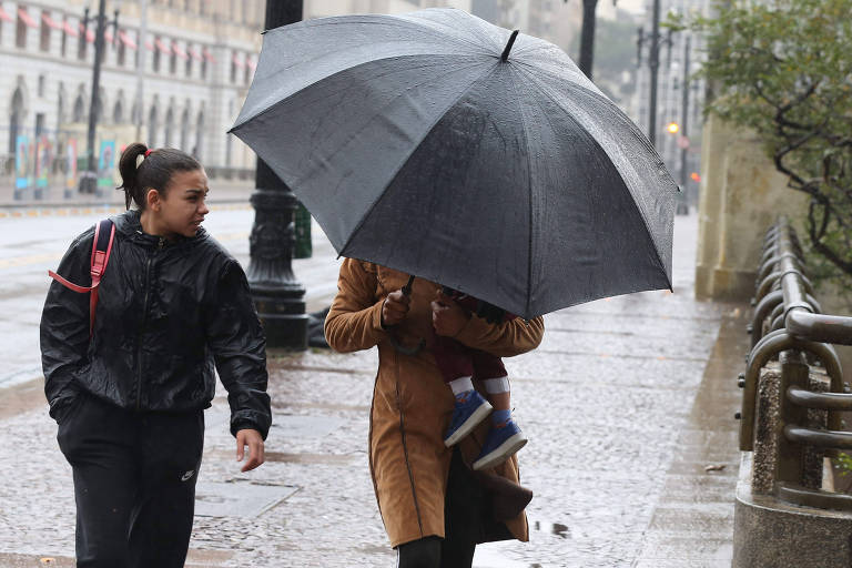 Duas mulheres caminham debaixo de chuva pelo Viaduto do Chá, em São Paulo. Uma delas está de jaqueta preta e a outra segura uma criança no colo e se protege debaixo de um guarda chuva preto.