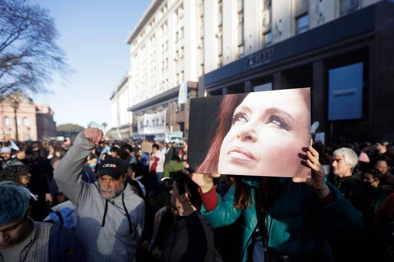 Pessoas fazem protesto em uma rua; uma delas ergue um retrato da vice-presidente argentina Cristina Kirchner tendo uma fachada de prédio ensolarada ao lado