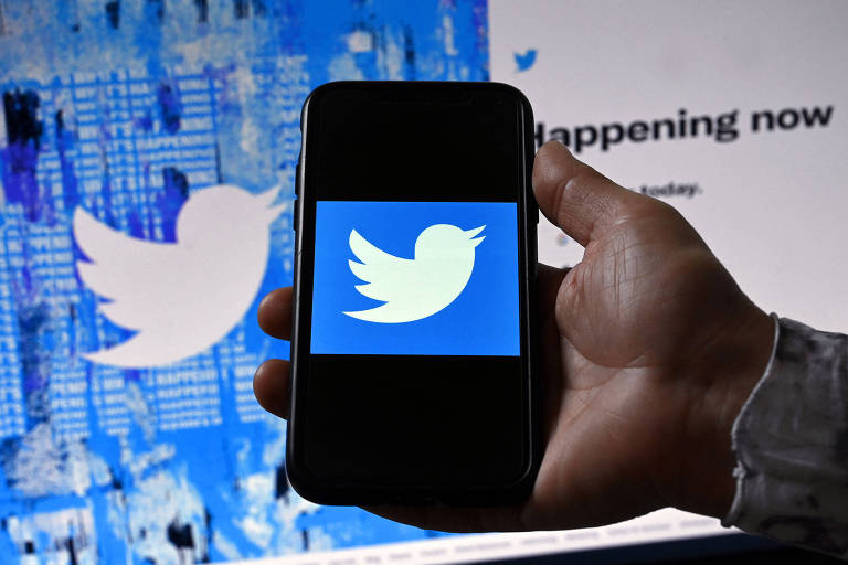 Imagem mostra mão segurando celular com logotipo do Twitter. Ao fundo, uma tela também mostra o logo.