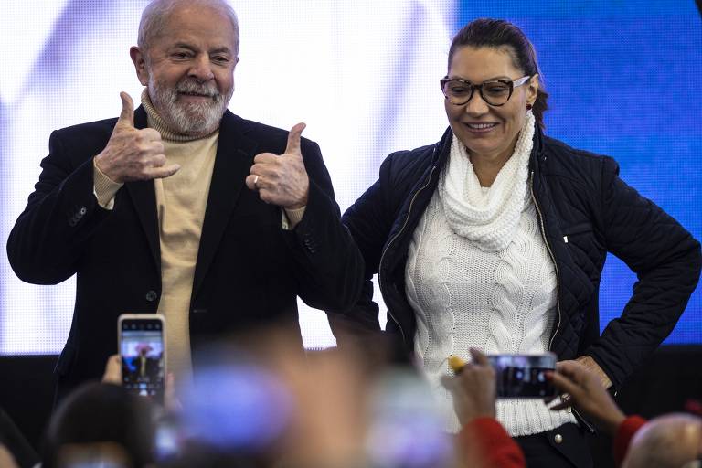 O ex-presidente Lula é um homem branco, idoso, com barba e cabelos grisalhos, orelhas de tamanho médio. Na foto está à esquerda, vestido com um blazer preto e uma camiseta bege. Ao lado dele está Janja, sua esposa. É branca, com o nariz um pouco achatado, usando óculos, com cabelo ondulado. Veste um blazer preto com uma camisa branca com bordados de oliveiras. Lula acena aos apoiadores, que estão na plateia com celulares apontados a ele, tirando fotos