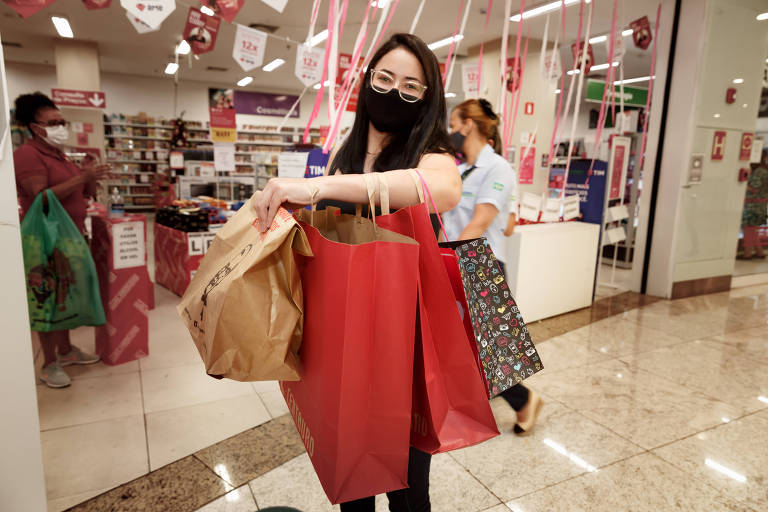 Mulher de óculos e máscara segura várias sacolas em frente a uma loja.