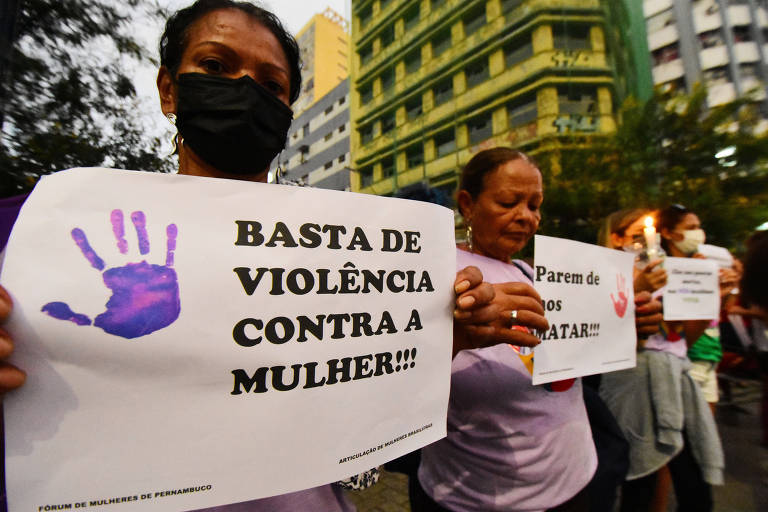 foto mostra mulher de máscara em protesto segurando cartaz com o texto "basta de violência contra a mulher". no cartaz há a palma de uma mão aberta na cor roxa