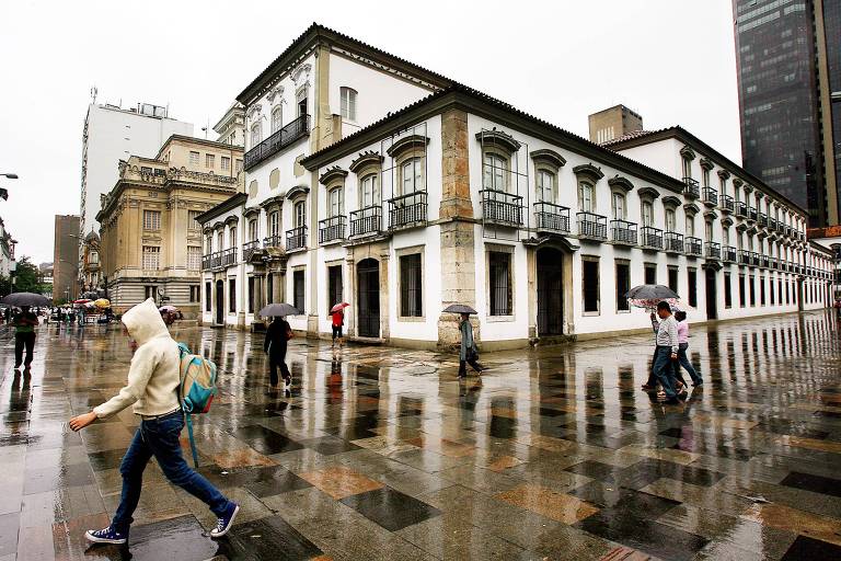 fachada do paço imperial num dia de chuva. alguns pedestres passam em frente ao espaço, com sombrinhas ou capuzes