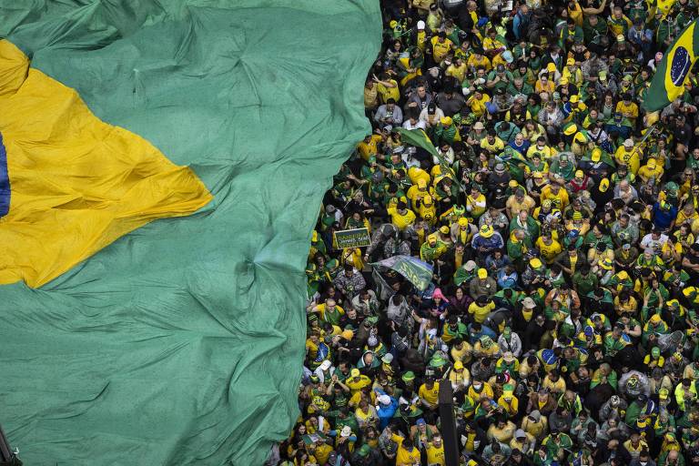 Imagem aérea mostra parte de bandeira do Brasil gigante encobrindo aglomeração de manifestantes. Predominância das cores verde e amarelo. 