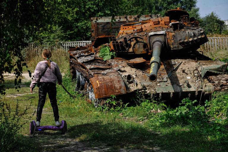 Menina ucraniana em um skate elétrico passa por tanque russo destruído em Lukachivka, norte do país