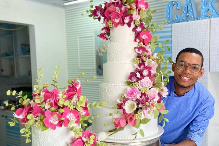 Homem negro usando óculos e dolma azul claro, sorrindo enquanto exibe um bolo de cinco andares decorado com flores vermelhas e rosas