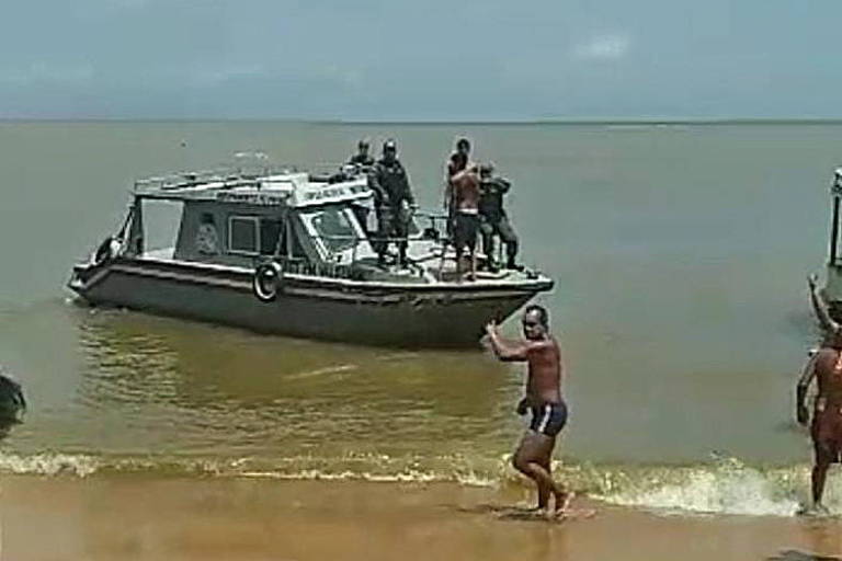 Barco com policiais navega perto de praia em busca de sobreviventes de naufrágio. Na areia da praia, um homem de sunga aponta em direção ao barco.