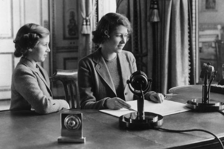 Foto em preto e branco mostra uma mulher mais velha e uma criança sentadas em frente a uma escrivaninha; as duas estão em frente a microfones