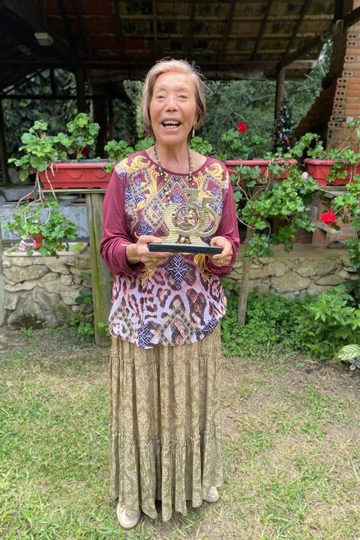Imagem colorida mostra a professora Hilda em pé, em um jardim; ela tem cabelo grisalho, usa uma roupa colorida em tons de vermelho e cinza; sorri e segura um troféu que tem o número 35 em dourado com as duas mãos.