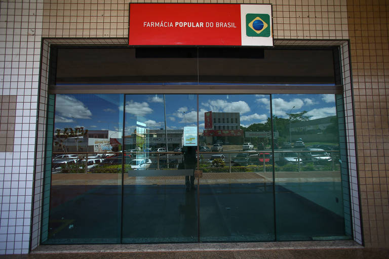 Imagem mostra fachada de farmácia. A porta é de vidro e, em cima dela, há uma placa vermelha com "Farmácia Popular" e uma bandeira do Brasil.