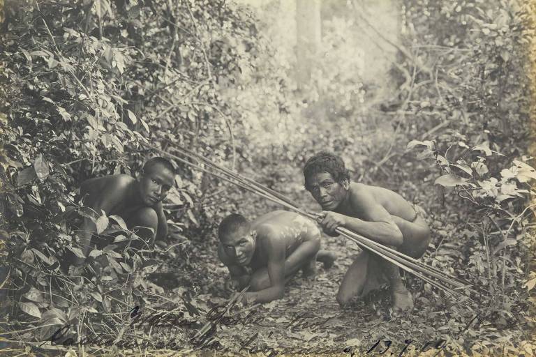 três homens indígenas são fotografados agachados no meio da mata. um deles aponta uma flecha para a câmera