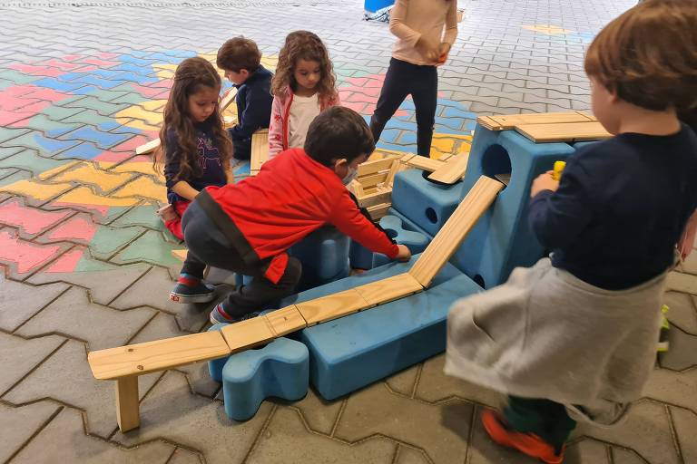 Na foto, seis crianças interagem com um brinquedo azul no centro da imagem. A base do brinquedo está no chão e sustenta uma rampa bege, de madeira, em que as crianças colocam uma bolinha para observar o movimento
