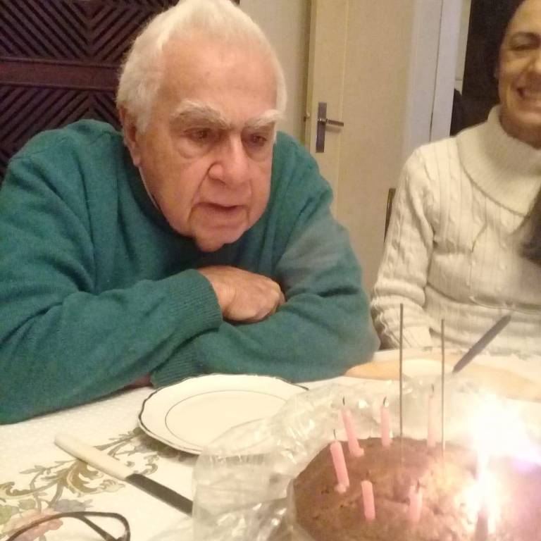 Foto de Carlos Eduardo Pellegrini Di Pietro. Ele era uma idoso, branco, de cabelos brancos e usava um suéter verde. Ele está sentado à mesa com um bolo de aniversário.