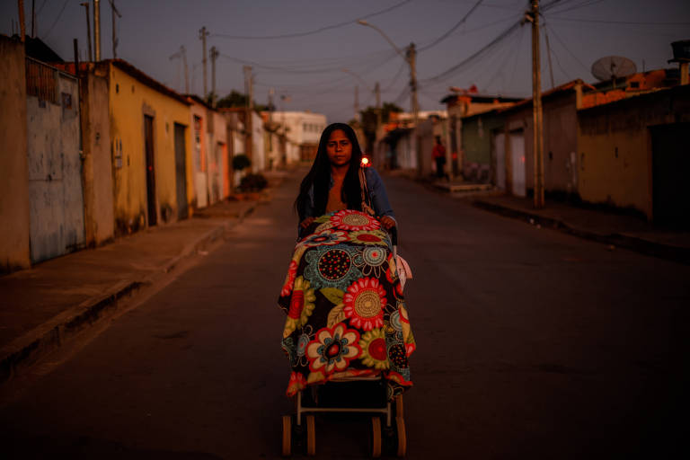 Thalia está de frente, com o carrinho de bebê à sua frente. O carrinho está coberto por uma manta colorida. ela está em uma rua com o céu ainda amanhecendo.