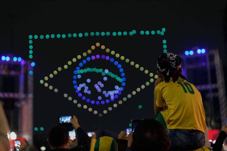 uma criança com a camisa do brasil, sentada sobre os ombros de uma pessoa na multidão, assiste ao show de drones à sua frente. em segundo plano, os drones formam a bandeira verde, amarela e azul do país