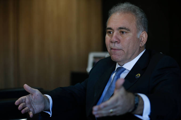 Foto mostra o ministro da Saúde Marcelo Queiroga em seu gabinete. Ele é um homem branco, que veste um terno preto, com camisa branca e gravata azul. Está sentado e gesticula.