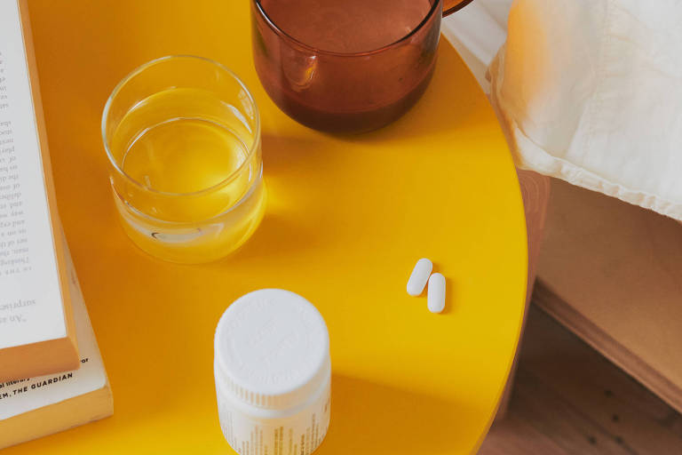 Xícara, copo, frasco de remédios e cápsulas ao lado da cama em um criado mudo amarelo. Foto tirada de cima
