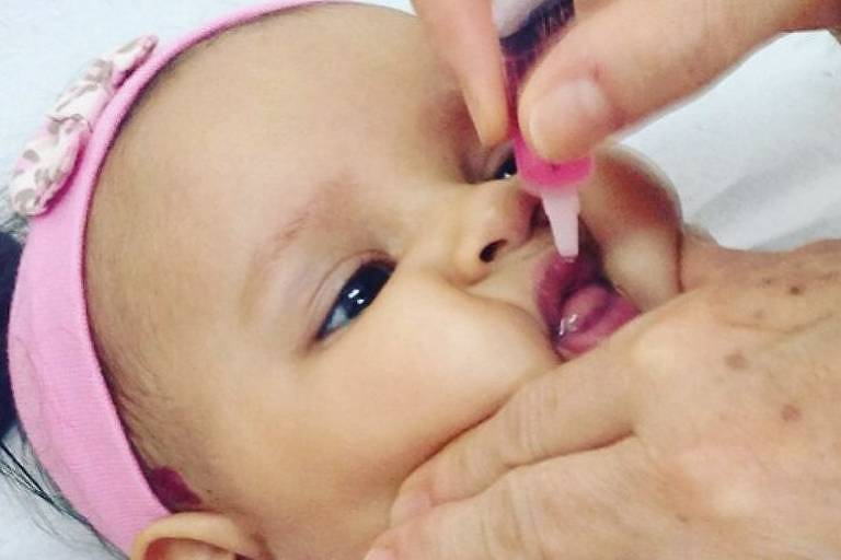 Um bebê com uma faixa cor de rosa nos cabelos recebe a gotinha da vacina contra a poliomielite. Na imagem é possível ver a mão da aplicadora, que usa um anel